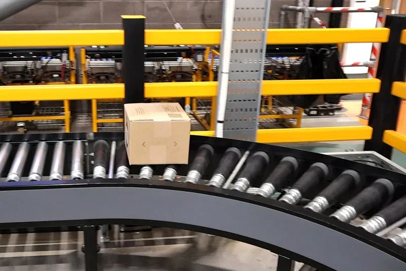 Ein Karton fährt um eine Kurve eines angetriebenen Rollenförderers mit gelben Sicherheitsbarrieren.